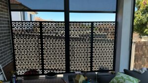 A large window with a decorative black perforated screen partially covering it, letting in sunlight. Outside are houses and a tree. Indoors, a green cushion and small items are visible near the window.