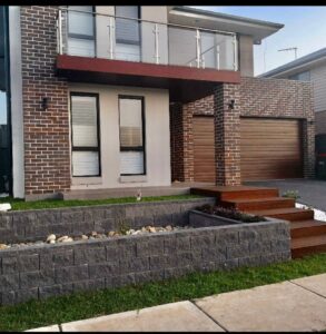 A modern two-story house with brown brick walls, large windows, a wooden balcony, a double garage, and stone-edged garden beds with rocks and plants along a lawn and wooden steps.