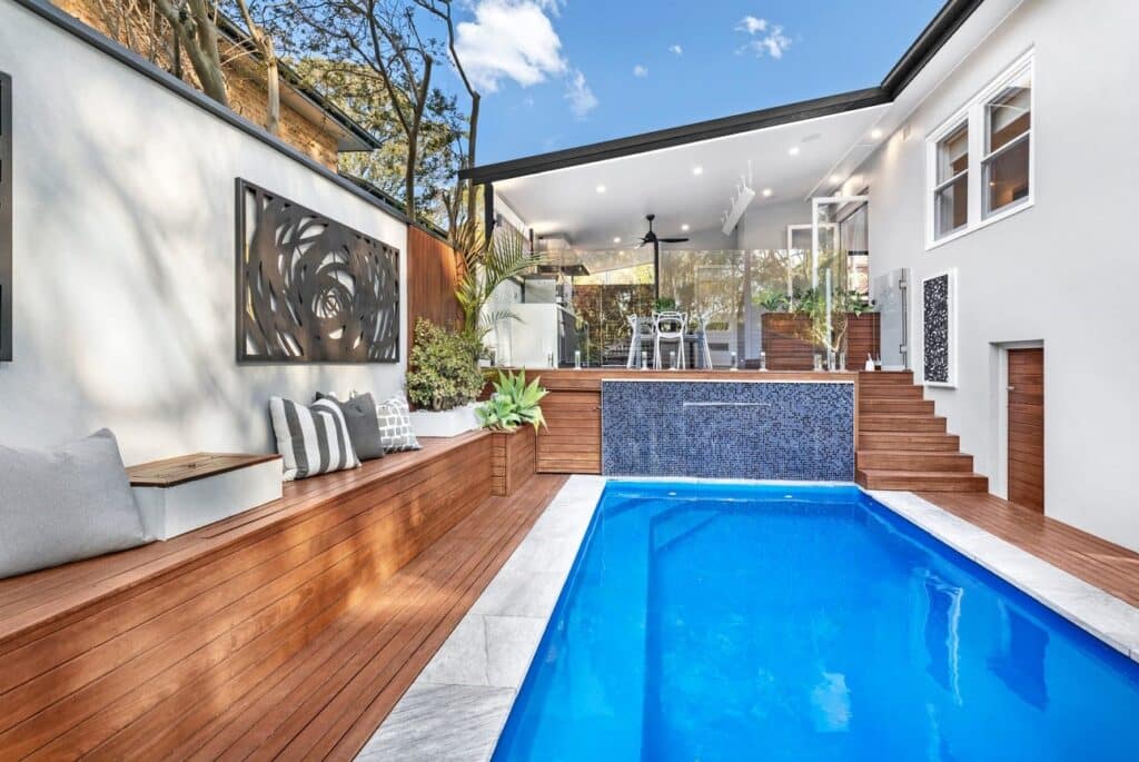 Modern backyard with a small rectangular pool, timber deck by a top decking service in Sydney, built-in bench with cushions, steps to a covered patio, outdoor dining area, and contemporary wall art. Trees and blue sky complete the scene.