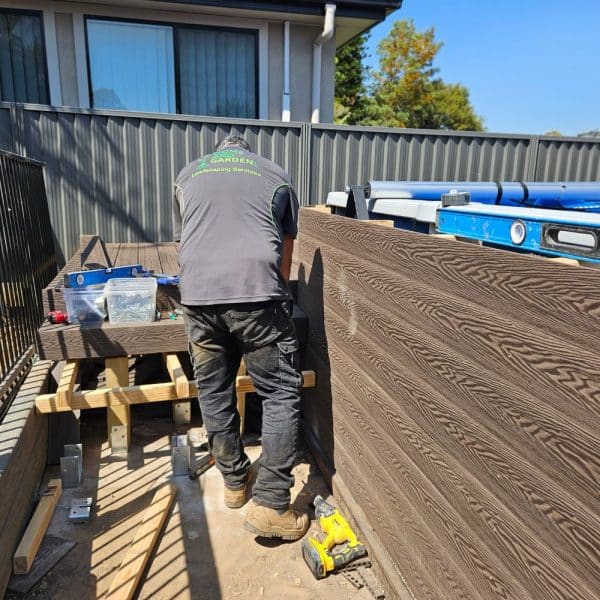 A worker in a gray shirt and black pants is building a wooden deck in a backyard, using tools and materials arranged around the work area near a gray fence and a house.the worker works for Home and Garden group