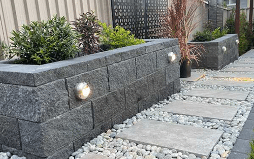A modern garden with raised stone planters, built-in round lights, potted plants, and a pathway made of square paving stones surrounded by small white pebbles.