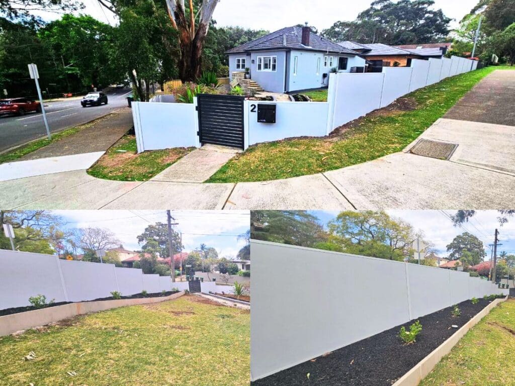 A modular wall white fence surrounds a corner house with a black gate, mailbox, and newly planted garden beds along the fence. The setting is suburban with lawns, trees, and sidewalks.