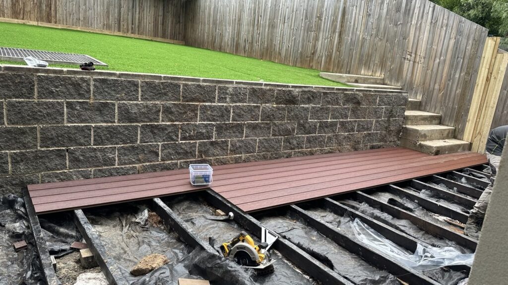A backyard renovation in progress shows a partially composite decking deck frame with brown decking boards being installed, tools scattered around, and artificial grass on a raised section bordered by a stone retaining wall.