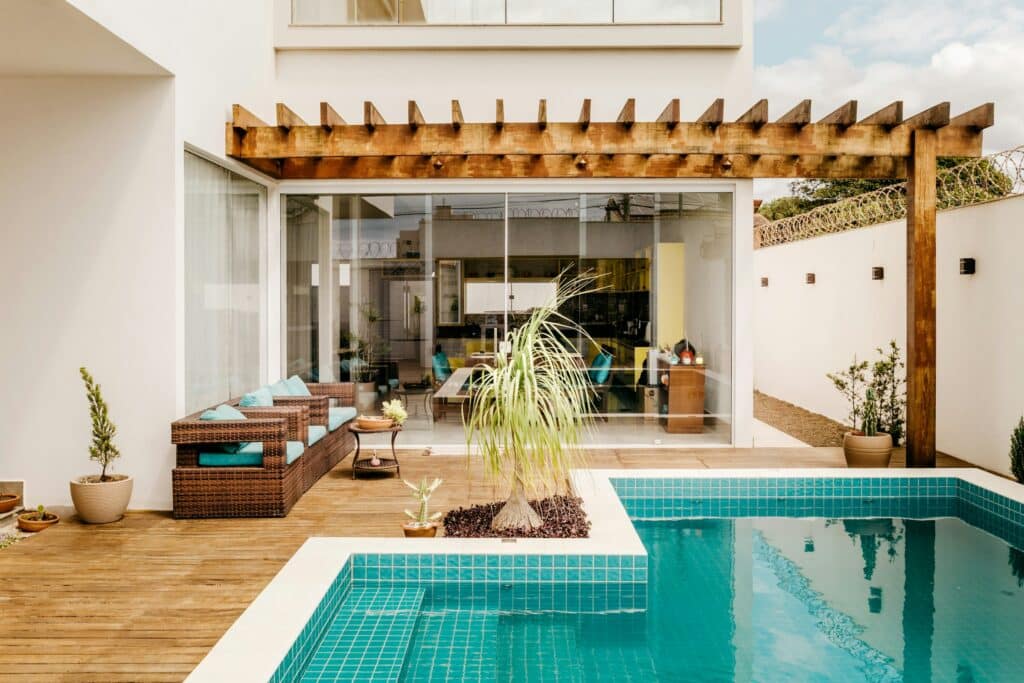 A modern backyard showcases expert outdoor landscaping in Sydney, with a turquoise-tiled pool, composite deck, wicker patio furniture with blue cushions, potted plants, and a wooden pergola beside a white house with large glass sliding doors.