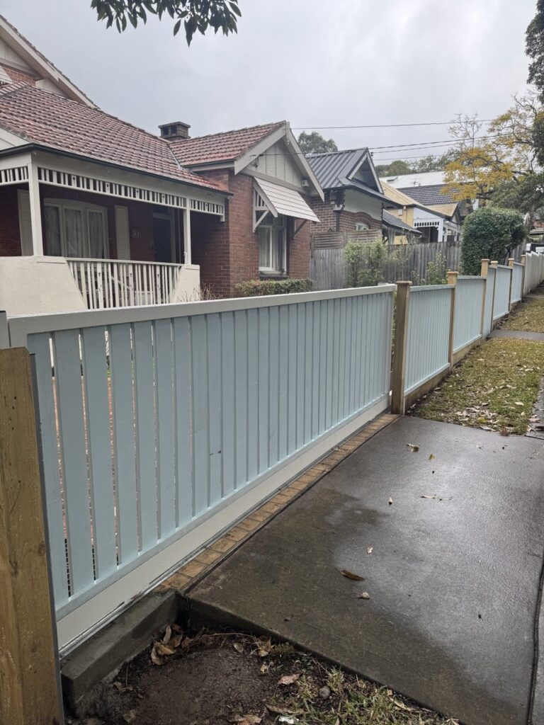 A light blue wooden fence runs along the sidewalk in front of brick houses with tiled roofs on a cloudy day. Wet pavement and scattered leaves indicate recent rain.