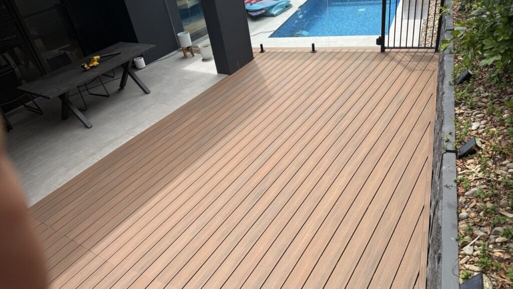 A modern outdoor deck with wood-like flooring next to a swimming pool, black metal fence, and a patio area with a table and chairs. Some plants and stones are visible along the right edge.