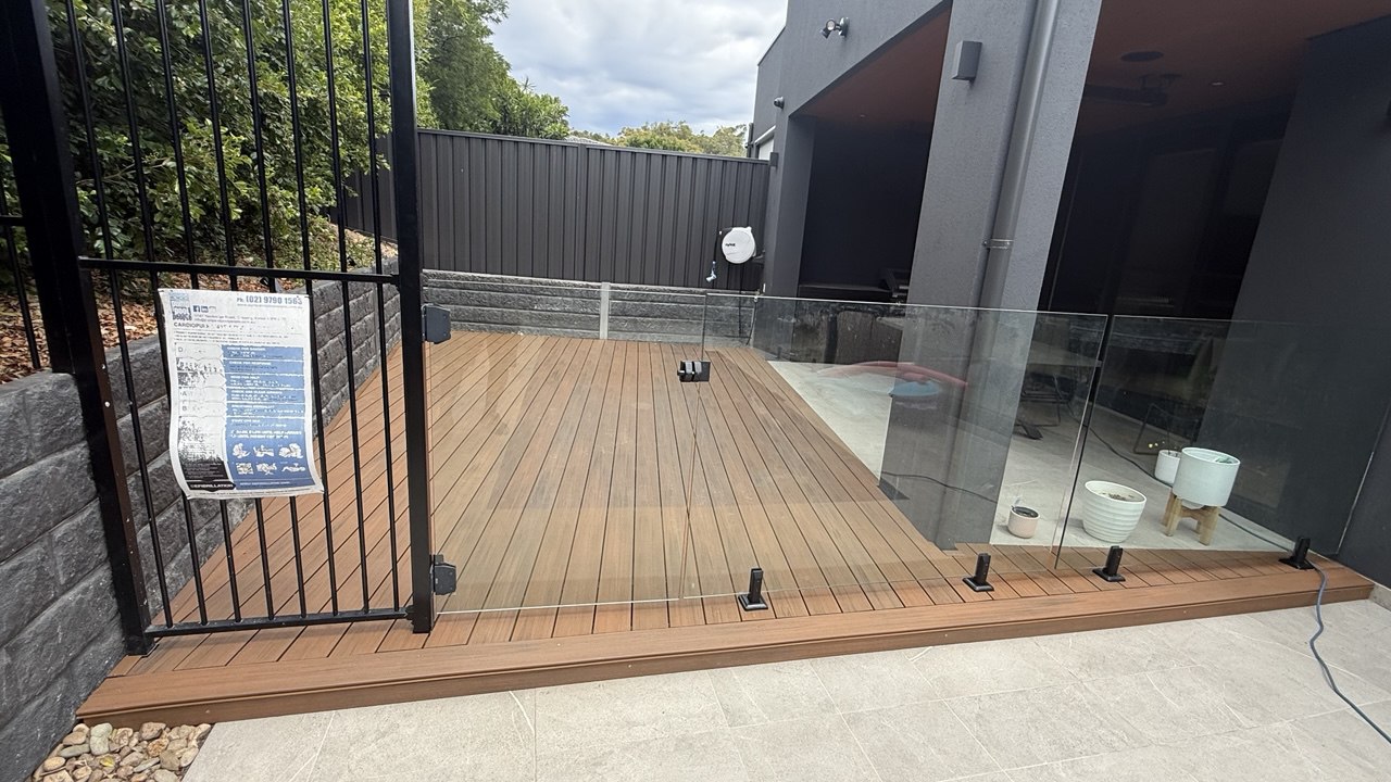 A fenced backyard area with a wooden deck enclosed by glass panels, next to a modern dark-colored house. There is a sign on the black metal gate and some outdoor items near the house wall.