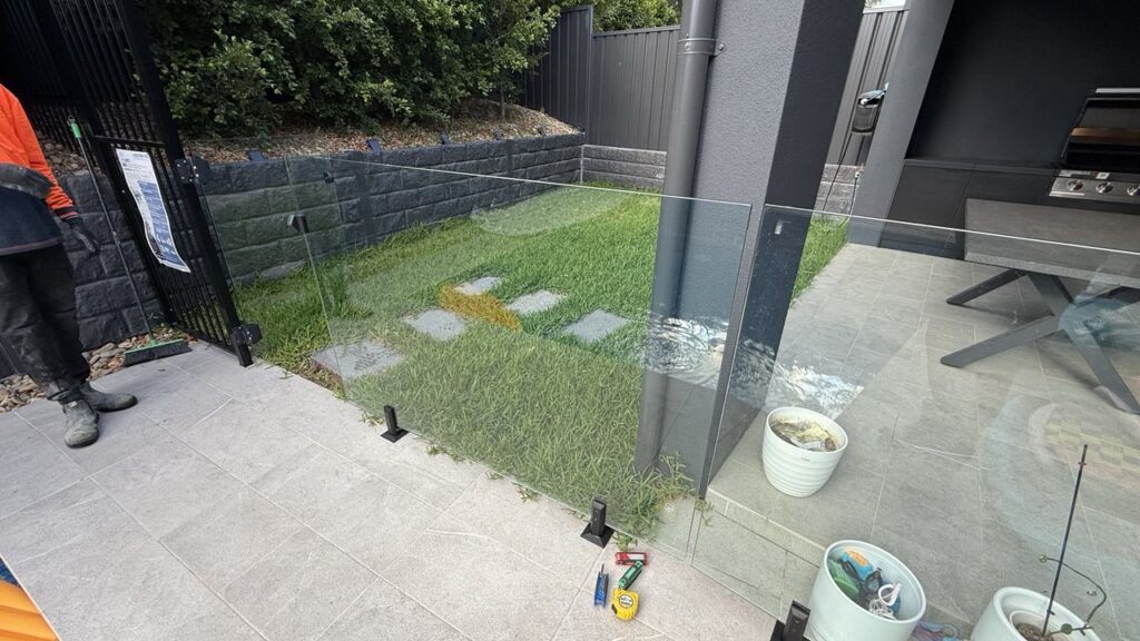 A glass fence separates a tiled patio with potted plants from a grassy yard with stepping stones. Tools are on the ground near the fence, and part of a person in an orange jacket is visible on the left.