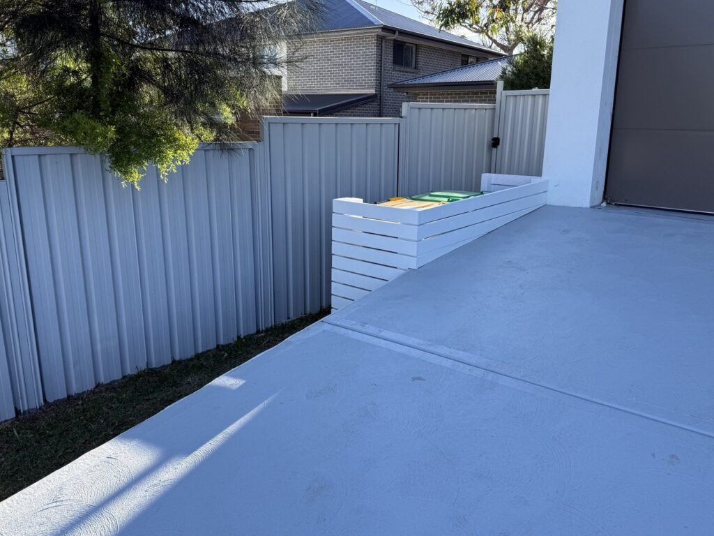 A sloped concrete driveway leads up to a modern house with a white planter box containing colorful bins beside a grey metal fence and green grass. Trees cast shadows onto the fence in the sunlight.