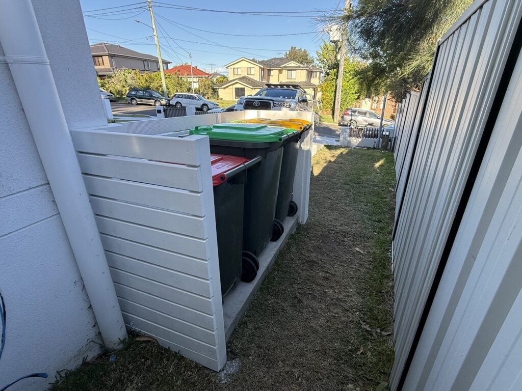 Four large outdoor bins with yellow, red, and green lids are lined up in a white wooden enclosure next to a fence, beside a grassy path near suburban houses and parked cars.
